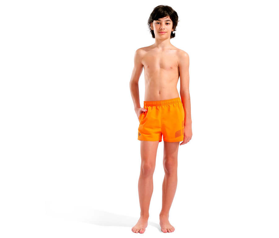 Un garçon pieds nus se tient devant un fond blanc et porte un short de bain orange.