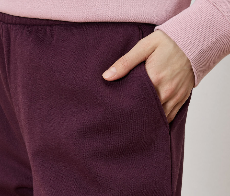 Gros plan d'une personne portant un pantalon de survêtement bordeaux et un pull rose, la main dans la poche.