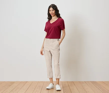 Une femme se tient devant un mur, portant un t-shirt rouge, un pantalon clair et des baskets blanches.