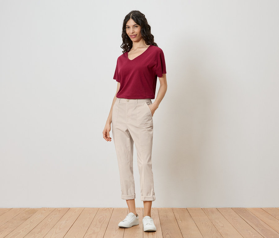 Une femme se tient devant un mur, portant un t-shirt rouge, un pantalon clair et des baskets blanches.