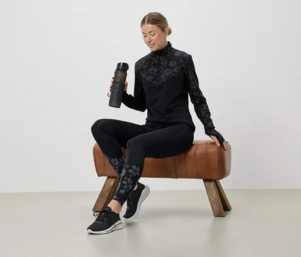 Une femme assise sur un cheval d'arçons tient une bouteille d'eau et porte une tenue de sport noire à motifs floraux.
