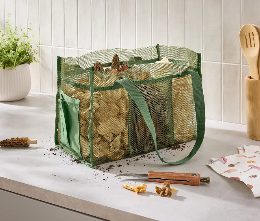 Sur un comptoir de cuisine, un sac en filet vert rempli de champignons est posé à côté d'un couteau, d'un torchon imprimé de champignons et d'un pot d'herbes aromatiques.