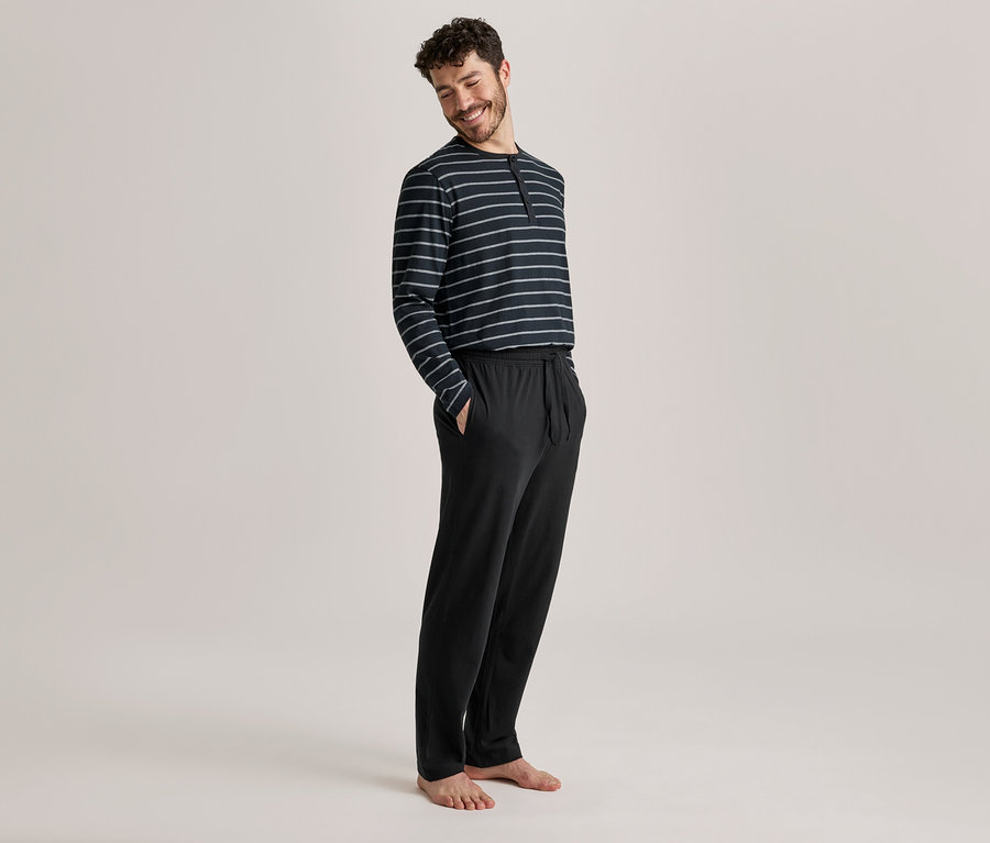 Homme souriant portant un haut à manches longues rayé foncé et un pantalon de pyjama noir, debout les mains dans les poches.