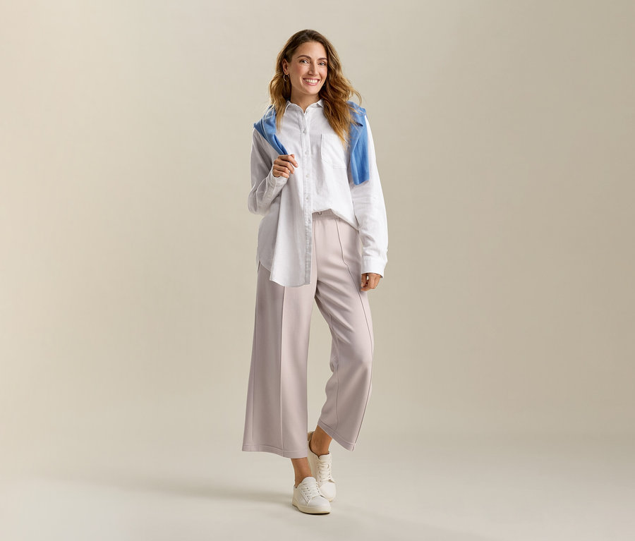 Une femme souriante porte une blouse longue en lin blanche et un pantalon en mélange de modal sable. Un pull bleu est posé sur ses épaules.