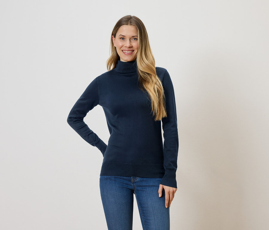 Une femme aux longs cheveux blonds portant un pull à col roulé bleu marine et un jean sur un fond blanc.