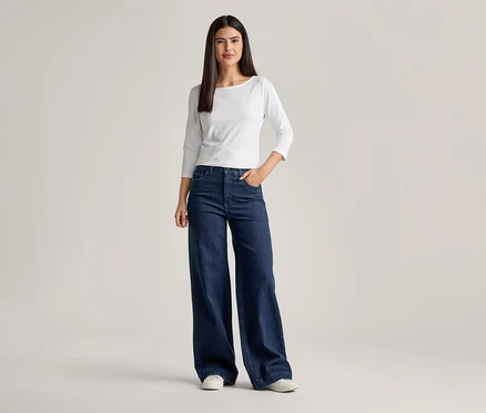 Femme portant un T-shirt blanc à manches longues et un jean évasé bleu foncé, une main dans la poche.