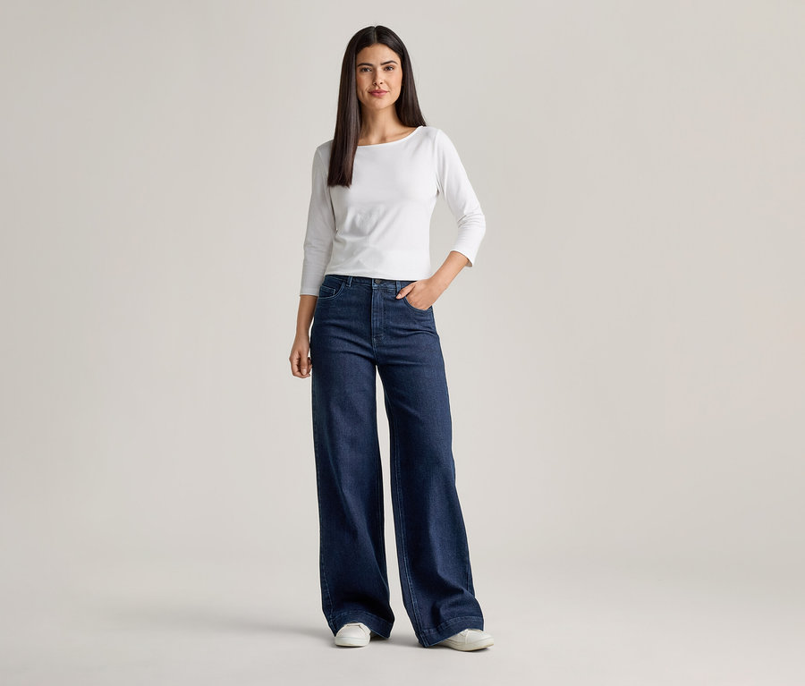 Femme portant un T-shirt blanc à manches longues et un jean évasé bleu foncé, une main dans la poche.