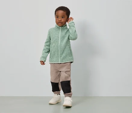 Un garçon se tient devant un mur blanc, vêtu d'une veste en polaire vert clair, d'un pantalon beige avec des empiècements noirs et de chaussures blanches.