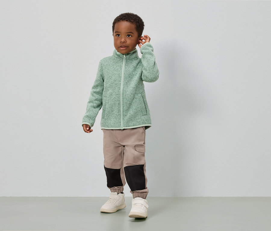 Un garçon se tient devant un mur blanc, vêtu d'une veste en polaire vert clair, d'un pantalon beige avec des empiècements noirs et de chaussures blanches.