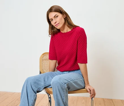 Une femme est assise sur une chaise, portant un haut rouge et un jean.