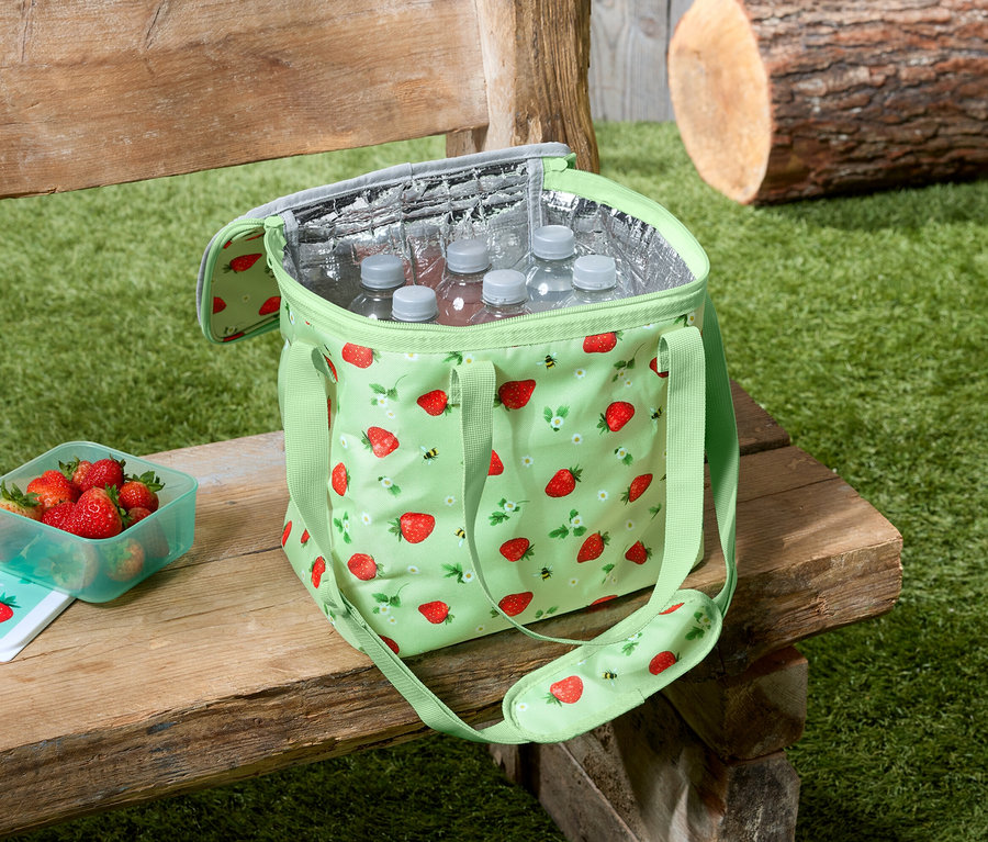 Un sac isotherme vert clair à motif fraises est posé sur un banc en bois, à côté d'un bol de fraises et des bouteilles d'eau dans le sac.