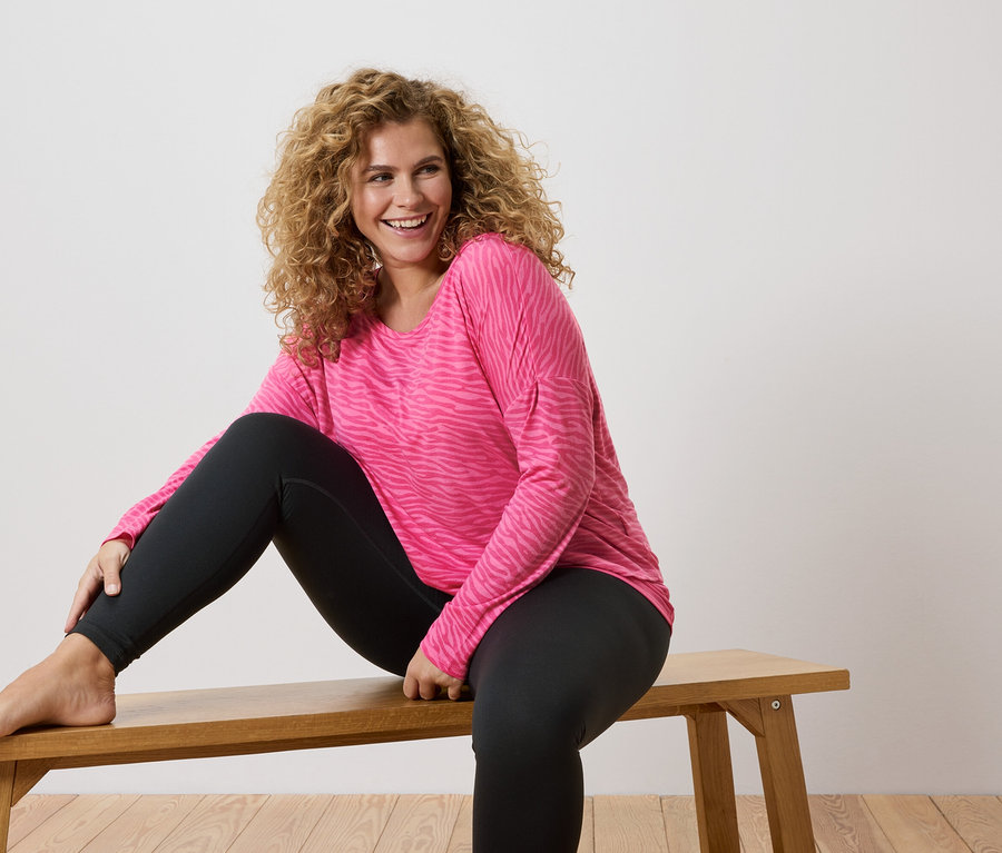 Une femme aux cheveux bouclés est assise pieds nus sur un banc en bois, portant un t-shirt rose à manches longues avec un motif zébré et un legging noir.