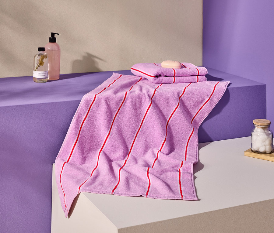 Photo de serviettes roses à rayures rouges, de savon, de bouteilles et de récipients sur une surface violette et crème.