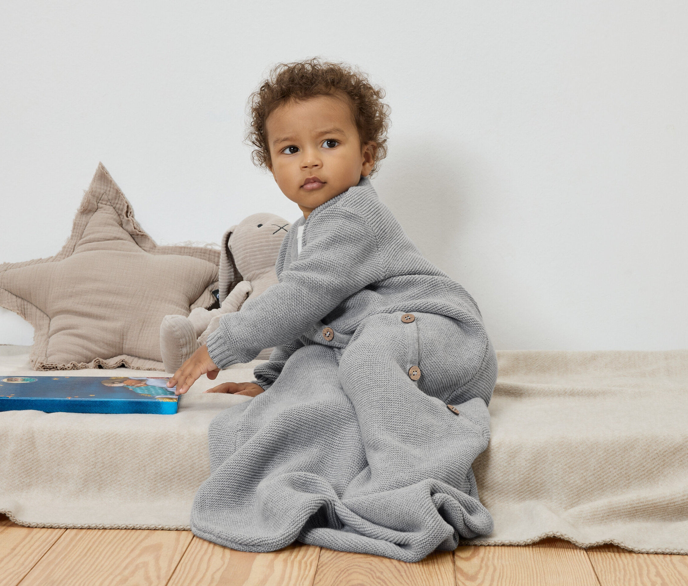 Bébé assis sur une couverture beige à côté d'une étoile et d'un lapin en peluche, touchant un livre bleu.