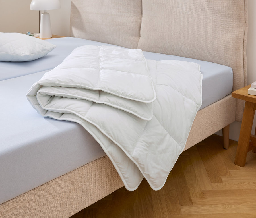 Une couette matelassée blanche Cell Solution® de taille normale est pliée sur un lit bleu clair à côté d'un oreiller.