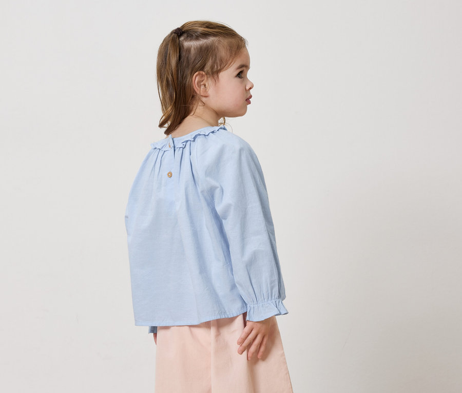 Fillette de dos, de côté, portant une chemise à volants bleue et un pantalon rose sur fond blanc.