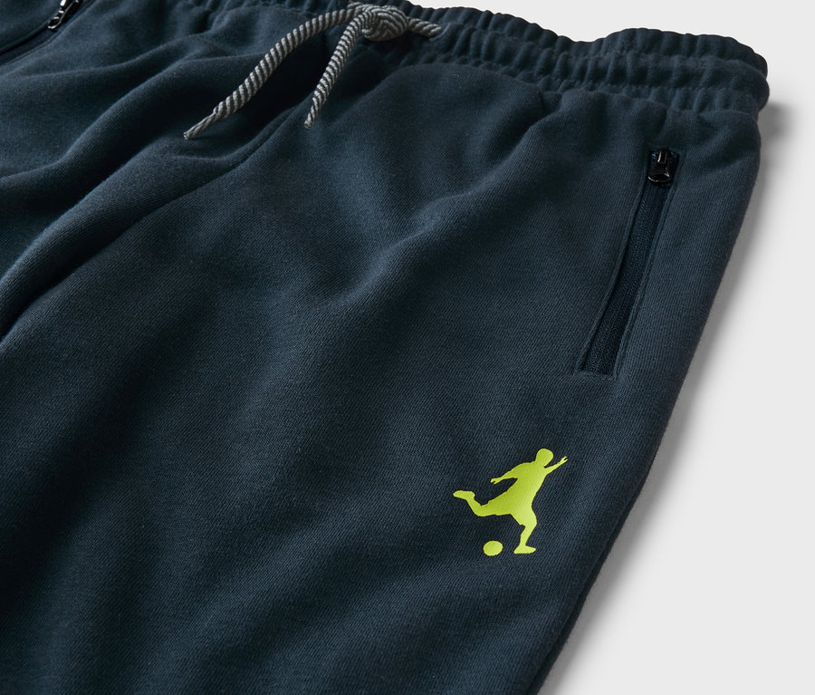 Gros plan d'un pantalon de survêtement bleu marine avec cordon de serrage, poche zippée et logo de joueur de football vert clair.