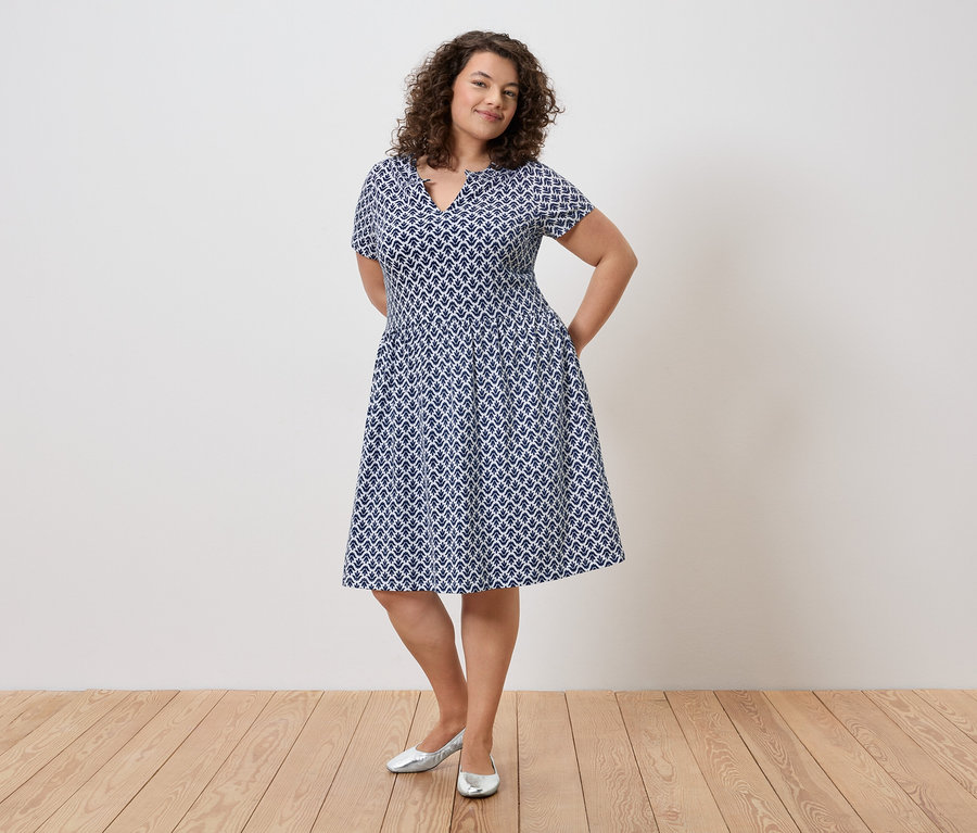 Femme aux cheveux bouclés portant une robe bleue et blanche et des chaussures argentées, les mains dans les poches, devant un mur.