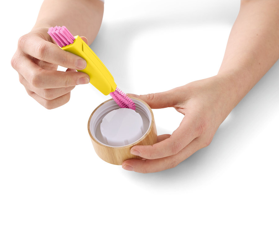 Une personne nettoie un couvercle en bambou avec une brosse de nettoyage jaune à poils roses.