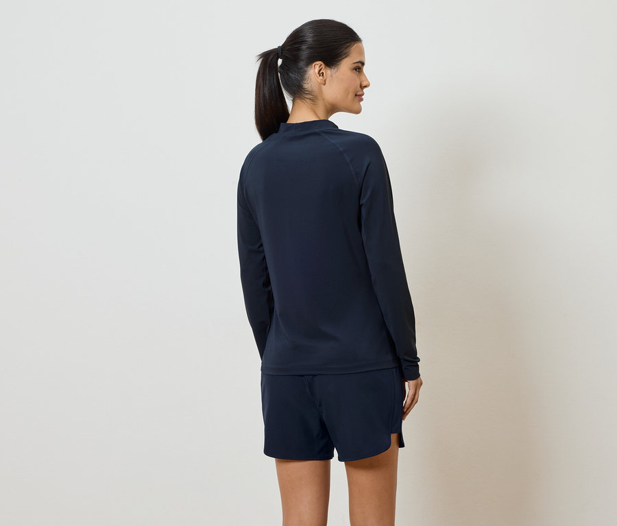 Une femme avec une queue de cheval porte un t-shirt à manches longues et un short bleu marine, vue de dos sur un fond crème.