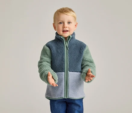 Un garçon blond aux yeux bleus porte une veste polaire bleue, verte et bleu clair, ainsi qu'un jean bleu.