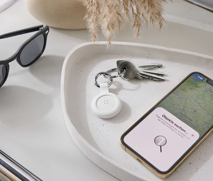 Sur un plateau reposent des lunettes de soleil, des clés, un traceur blanc et un smartphone avec une application de carte.