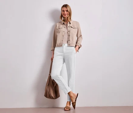 Une femme souriante est debout devant un mur blanc, vêtue d'une veste en jean beige, d'un haut clair et d'un pantalon chino blanc Street One. Elle tient un sac à bandoulière marron et porte des sandales marron.