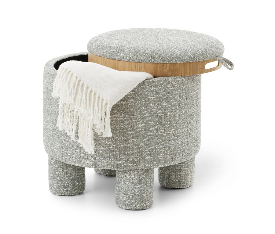 Pouf rond gris rembourré avec quatre pieds courts, un couvercle-plateau en bois partiellement ouvert et un plaid blanc à franges drapé sur le côté.