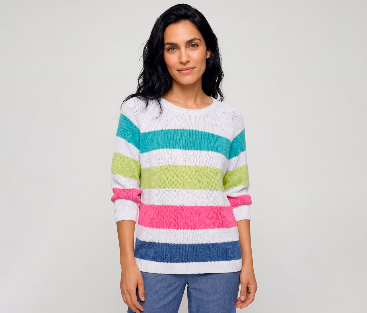 Une femme porte un pull Olsen rayé blanc avec des rayures turquoise, vert anis, rose vif et bleu, et un pantalon bleu.