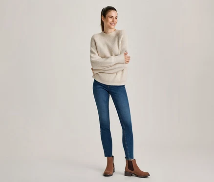 Femme avec une queue de cheval, souriante et regardant à droite, portant un pull côtelé crème, un jean skinny bleu et des bottines marron.