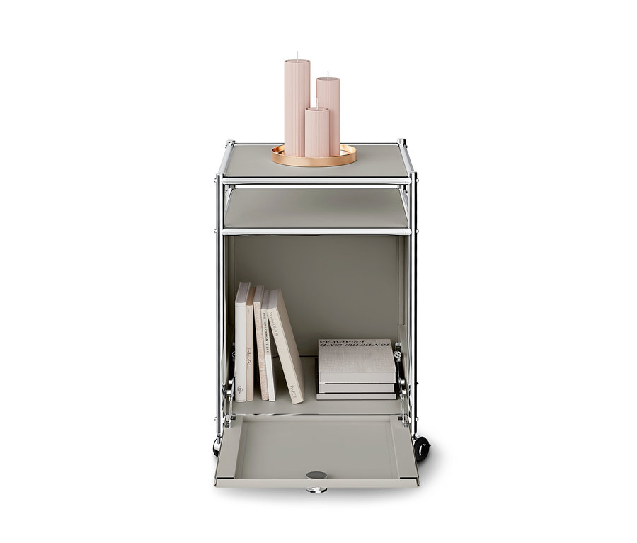 Meuble modulaire gris à roulettes avec un cadre chromé. Sur le dessus, un plateau doré contient trois bougies roses côtelées. Le compartiment inférieur est ouvert, révélant plusieurs livres, certains debout et d'autres empilés.