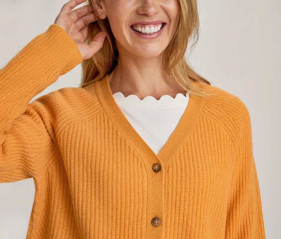 Une femme souriante porte un cardigan orange côtelé sur un haut blanc à col festonné, une main près de son oreille.