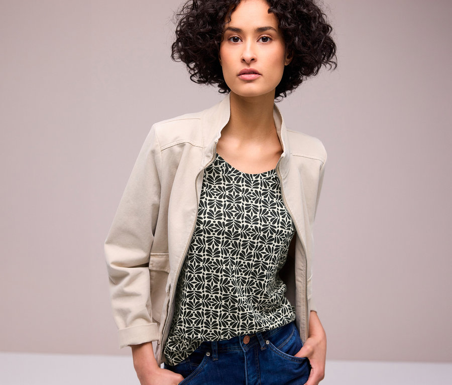 Une femme aux cheveux bouclés porte une veste beige ouverte sur un T-shirt Street One à motifs et un jean bleu, les mains dans les poches.