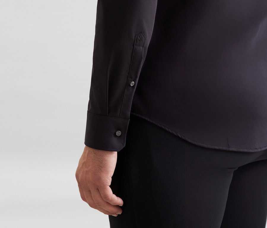 Personne portant une chemise noire à manches longues avec des boutons aux poignets et un petit détail brodé sur la manche, associée à un pantalon noir.
