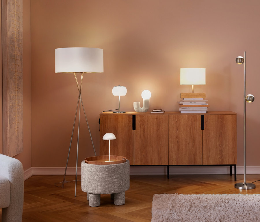Un salon avec plusieurs lampes allumées. Une lampe de table à LED sans fil blanche est posée sur une table d'appoint ronde, à côté d'un fauteuil et d'un buffet en bois avec d'autres lampes.