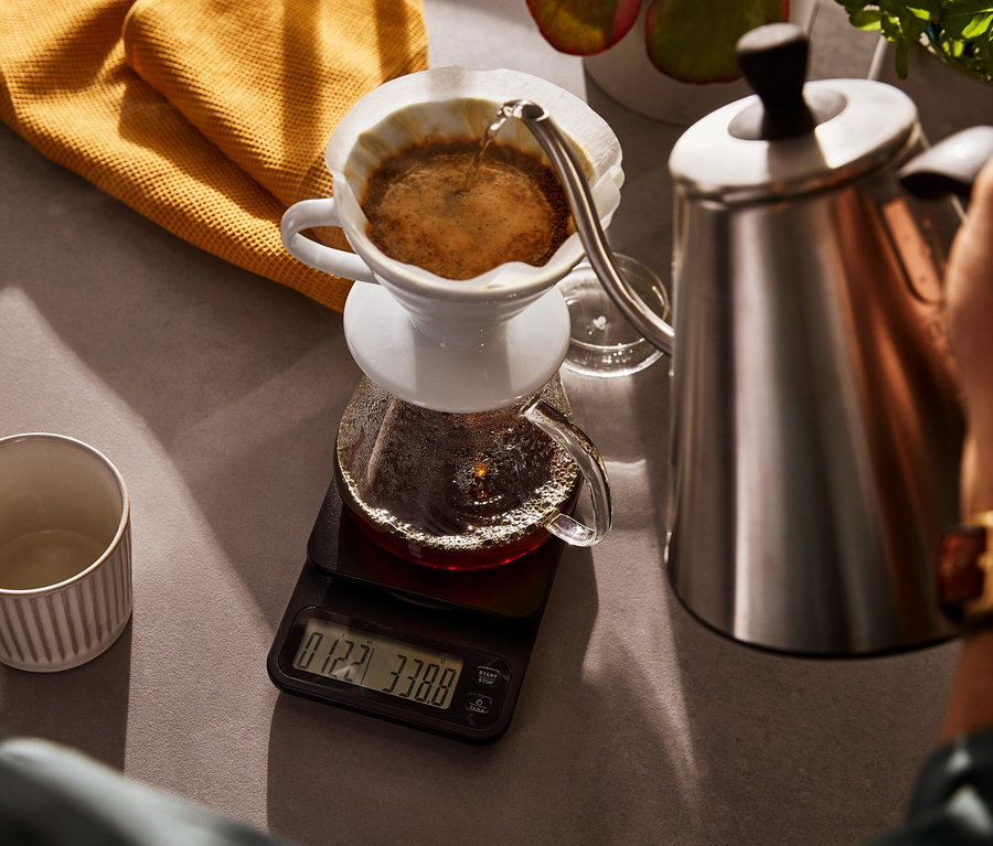 Une personne verse de l'eau d'une bouilloire électrique à col de cygne dans une cafetière en verre, laquelle repose sur une balance à café avec minuterie.