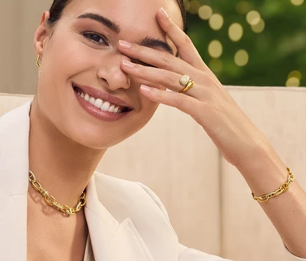 Une femme est assise sur un canapé, souriant et se couvrant un œil avec sa main. Elle porte une veste, un collier et un bracelet.