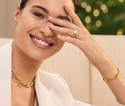 Une femme est assise sur un canapé, souriant et se couvrant un œil avec sa main. Elle porte une veste, un collier et un bracelet.