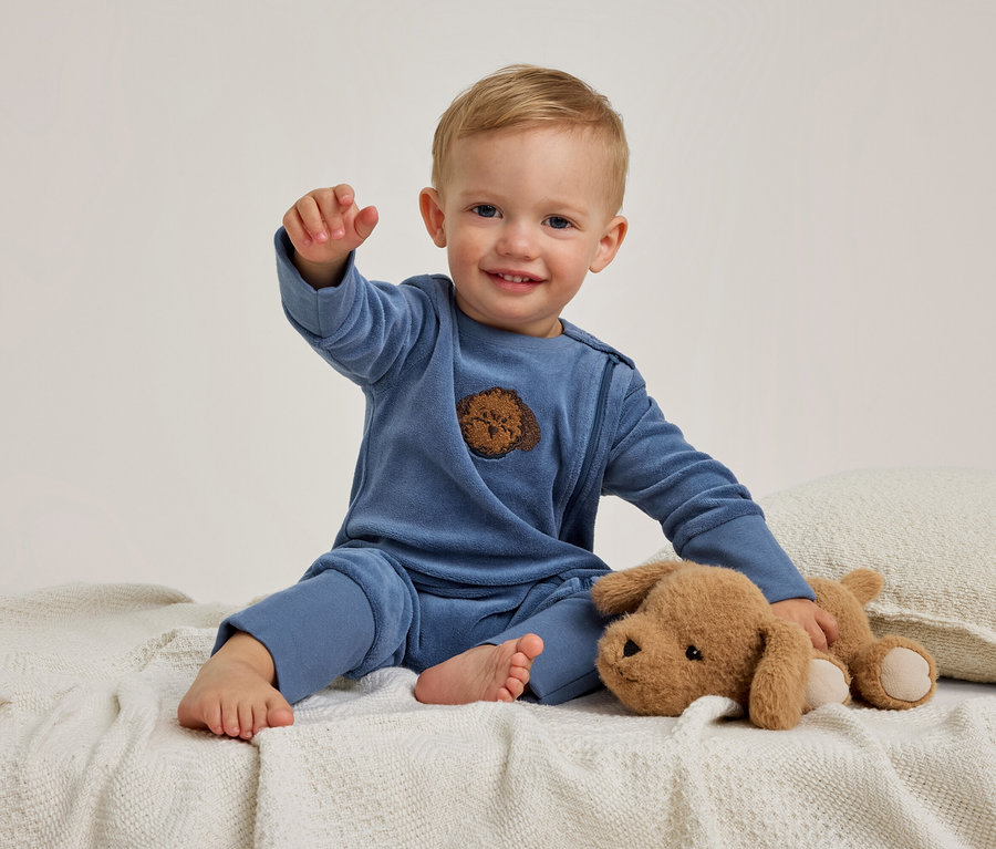 Un bébé souriant en barboteuse bleue est assis sur une couverture blanche, tendant une main. Un chien en peluche marron est allongé à côté du bébé.