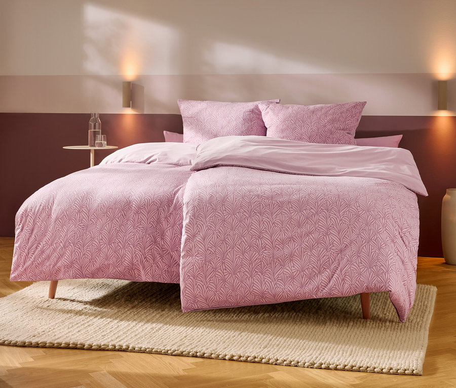 Un lit avec une parure de lit rose à motifs et des oreillers, ainsi que des draps roses unis, est posé sur un tapis clair sur un parquet en bois. Une petite table ronde avec une bouteille d'eau et un verre se trouve à gauche. Deux appliques murales éclairent le mur rose bicolore derrière le lit, et un grand vase est à droite.