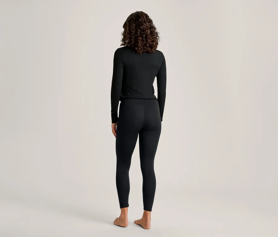 Une personne aux cheveux foncés et bouclés se tient dos au spectateur, vêtue d'un haut noir à manches longues et d'un legging noir. Elle est pieds nus devant un fond clair et uni.