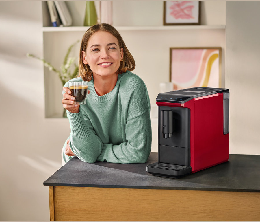 Femme souriante en pull vert clair tenant un verre de café, appuyée sur un comptoir sombre à côté d'une machine à café rouge et noire.