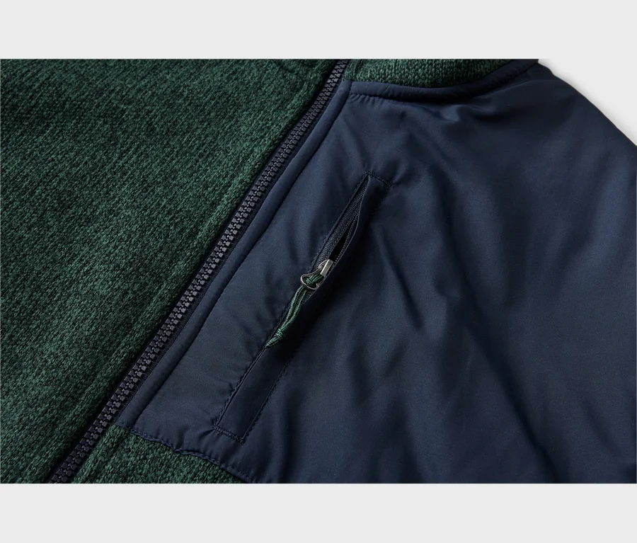 Gros plan d'un vêtement avec un tissu tricoté vert foncé à gauche et un tissu lisse bleu foncé à droite, séparés par une fermeture éclair noire. Une poche zippée avec une tirette verte est sur la section bleue.