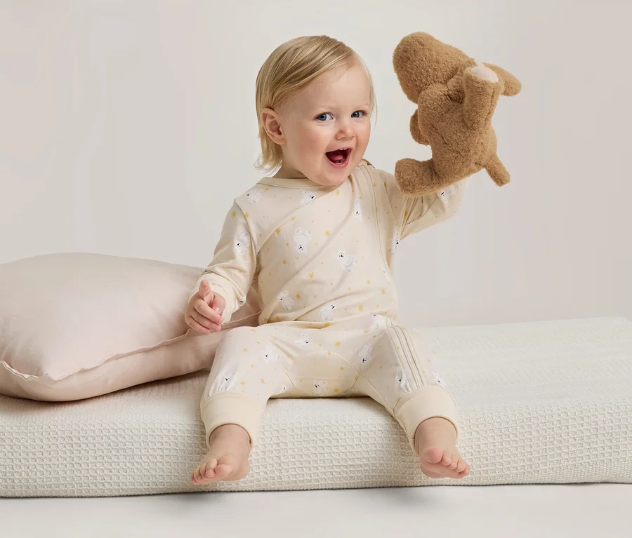 Un bébé souriant en pyjama jaune clair à motifs de moutons est assis sur une couverture blanche, tenant un animal en peluche marron. Un oreiller rose est à côté.