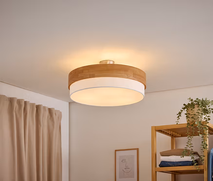 Un plafonnier rond avec un dessus en bois et un diffuseur blanc éclaire une pièce. Un rideau beige est à gauche, et une étagère en bois avec des vêtements et une plante est à droite.