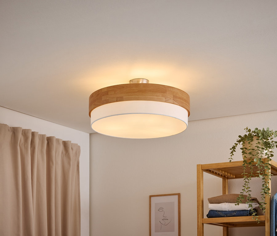 Un plafonnier rond avec un dessus en bois et un diffuseur blanc éclaire une pièce. Un rideau beige est à gauche, et une étagère en bois avec des vêtements et une plante est à droite.