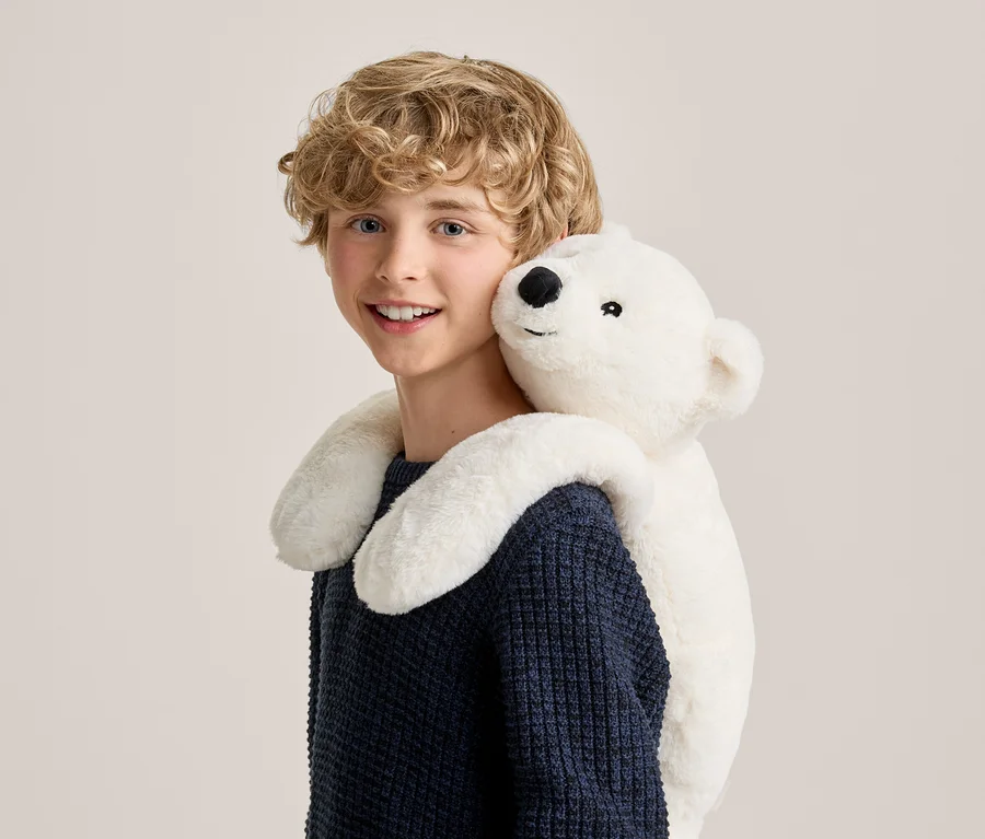 Garçon souriant aux cheveux blonds bouclés portant un pull bleu foncé, étreint par derrière par une peluche ours polaire blanche drapée sur ses épaules.