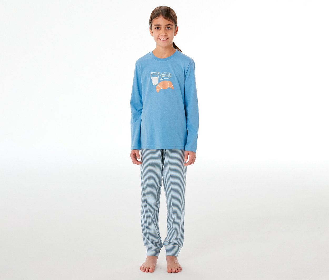 Fille souriante portant un pyjama. Le haut bleu clair à manches longues est imprimé d'un verre de lait, d'un croissant et du mot "Bonjour". Le pantalon est rayé bleu clair et blanc.