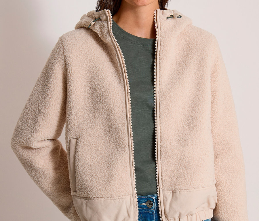 Une femme souriante porte une veste CECIL beige en polaire teddy à capuche, un t-shirt vert foncé et un jean bleu.