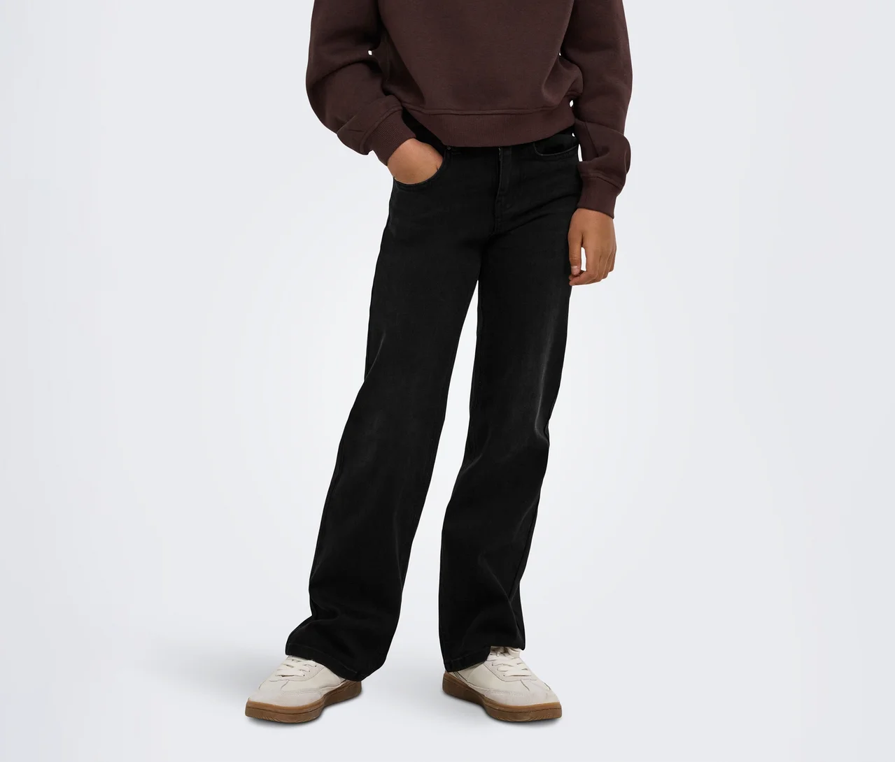 Personne portant un sweat-shirt marron, un jean noir large avec une main dans la poche et des baskets beige clair.